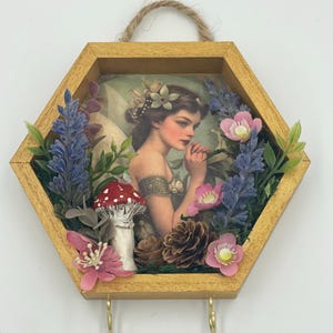 May include: A hexagonal wooden frame with a gold finish, featuring a vintage illustration of a fairy with wings, surrounded by artificial flowers, leaves, and a red and white mushroom. Two gold hooks are attached to the bottom of the frame.