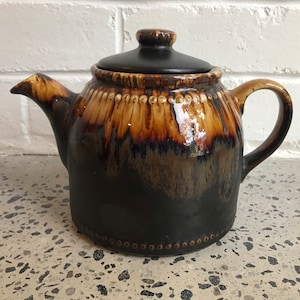 Może przedstawiać: Czarno-brązowy ceramiczny czajnik z pokrywką. Czajnik ma brązową glazurę z czarnym efektem kapania. Czajnik ma okrągły korpus i dzióbek po boku.