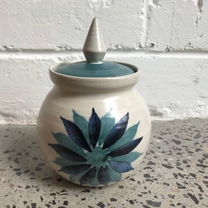 May include: A white ceramic jar with a blue and green floral design and a white pointed lid.