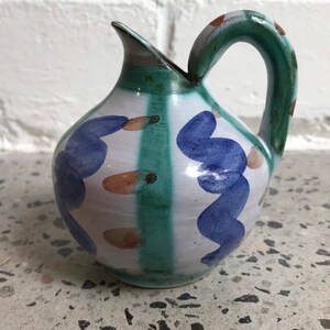 Può includere: Un piccolo brocca in ceramica bianca con manico verde e disegni dipinti in blu e marrone.