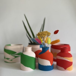 May include: Four small ceramic vases with abstract designs in shades of green, red, blue, and white. The vases are arranged in a row on a white surface. Dried flowers are in one of the vases.