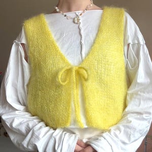 May include: A bright yellow sleeveless knit vest with a fuzzy texture and a V-neckline. The vest features a thin matching yellow tie closure at the center front. It is layered over a long-sleeved white shirt with visible shoulder cutouts.