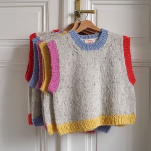 Könnte beinhalten: Eine Reihe farbenfroher Strickwesten, die an einem Holzkleiderbügel hängen. Die Westen sind grau mit Sprenkeln und haben jeweils einen farbigen Besatz: rot, blau, gelb und rosa. Sie hängen an einer weißen Tür.