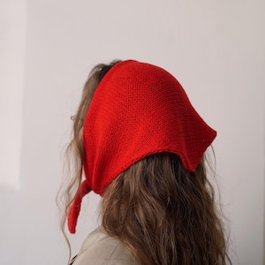 Può includere: Una sciarpa rossa brillante a maglia, annodata sulla nuca. Il copricapo è realizzato in un tessuto a maglia strutturato. La persona indossa una giacca beige. Lo sfondo è un muro bianco liscio.