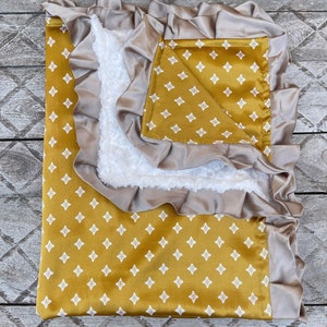 May include: A mustard yellow baby blanket with a white diamond pattern. The blanket has a satin trim and a soft, white sherpa lining.