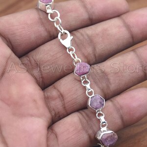 Può includere: Un braccialetto d'argento con una serie di pietre preziose rosa di forma irregolare. Le pietre sono collegate da una delicata catena e chiusura in argento. Il braccialetto è esposto su una mano.