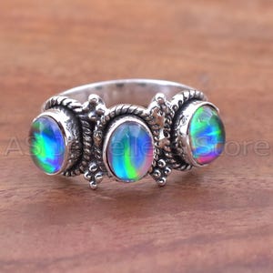 May include: A silver ring featuring three oval-shaped, iridescent gemstones. The gemstones display a spectrum of colors, including blue, green, and purple. The ring has a detailed, textured band with decorative elements. The ring is on a wooden surface.