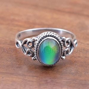 May include: Silver ring with an oval, color-changing stone. The stone shifts from blue to green. The ring has a decorative, openwork design on the sides and a twisted rope border around the stone. The text "AS Jewellers Store" is visible.