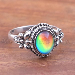 May include: Silver ring with an oval, color-shifting gemstone. The gemstone displays a spectrum of colors, including red, yellow, green, and blue. The ring has a detailed, ornate design around the gemstone.