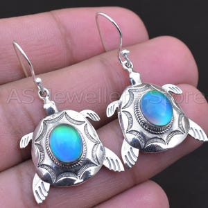 May include: A pair of silver-toned turtle earrings. Each earring features a turtle design with a central oval-shaped iridescent stone that shifts between blue and green hues. The turtles have detailed flippers and textured shells.