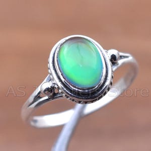 May include: A silver ring featuring an oval-shaped, iridescent gemstone with green and blue hues. The ring has a detailed, textured setting and a delicate band. The gemstone appears to shift colors, creating a unique and eye-catching piece of jewelry.