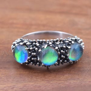 May include: A silver ring featuring three iridescent, oval-shaped stones. The stones display a range of colors, including blue, green, and yellow. The band has a textured, ornate design. The ring is set against a wooden surface.