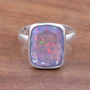 May include: A silver ring featuring a large, rectangular, faceted opal gemstone. The opal displays a play of color with flashes of red, green, and blue within a lavender base. The ring is set on a wooden surface.