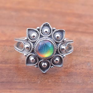May include: A silver-colored ring with a central iridescent stone. The ring features a floral design with intricate detailing around the stone and along the band. The ring is displayed on a wooden surface.