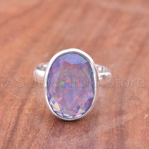 May include: A silver ring with a large, oval-cut, iridescent gemstone. The gemstone displays a range of colors, including purple, blue, and green. The ring is set against a wooden background, with the text "AS Jeweller Store" visible.