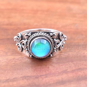 May include: A silver ring with a round, iridescent blue-green stone at the center. The ring has a detailed, ornate design with swirling patterns and a twisted rope-like border around the stone. The ring is set against a wooden surface.
