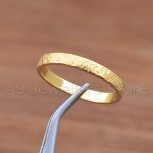 Puede incluir: Un anillo de oro con una superficie texturizada. El anillo está sujeto por unas pinzas plateadas sobre un fondo marrón. El anillo tiene un diseño sencillo y elegante.