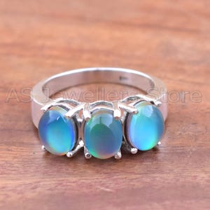 May include: A silver ring featuring three oval-cut, iridescent gemstones. The stones display a blend of blue and green hues. The ring is set against a wooden surface, with the text "AS Jeweller Store" visible.