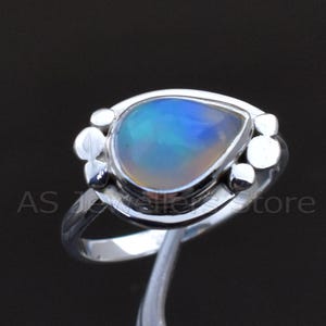 May include: A silver ring featuring a pear-shaped opal gemstone with blue, green, and yellow hues. The ring has a decorative setting with small, round accents. The ring is displayed against a dark background.