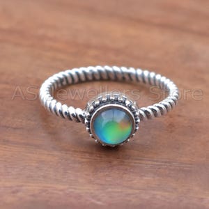 May include: A silver ring with a twisted band and a central, round, iridescent stone. The stone displays a spectrum of colors, including green, blue, and yellow. The ring is set against a wooden surface.