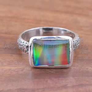 May include: A silver ring with a rectangular, iridescent gemstone. The gemstone displays a spectrum of colors, including green, red, and blue. The band has a textured design. The ring is on a wooden surface.