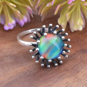 May include: A silver ring featuring a large, iridescent, round stone with a rainbow of colors. The stone is surrounded by black metal spikes tipped with small silver beads. The ring is set against a wooden surface.