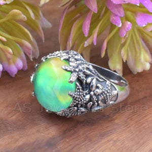 May include: A silver ring with a large, oval-shaped, iridescent green gemstone. The ring's setting features intricate starfish and sea creature designs. The ring is displayed on a wooden surface, with blurred flowers in the background.