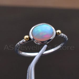 May include: A silver ring with an oval opal gemstone. The opal has a mix of blue, pink, and white colors. The ring has two small gold-colored beads on either side of the opal. The band has a textured design.