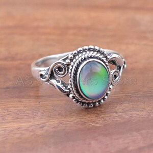 May include: A silver ring with an oval, iridescent stone. The stone shifts colors, displaying hues of green and purple. The ring has a detailed, decorative setting with a rope-like border and scrollwork accents. The text "AS Jewelers Store" is visible.