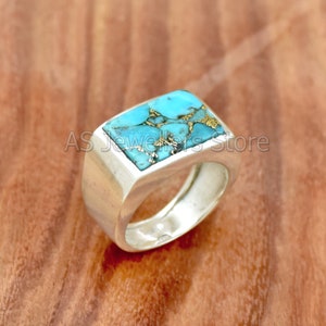 以下が含まれることがあります： 中央に長方形のターコイズストーンがセットされたシルバーリング。ストーンは青緑色で、全体に金色の斑点がある。