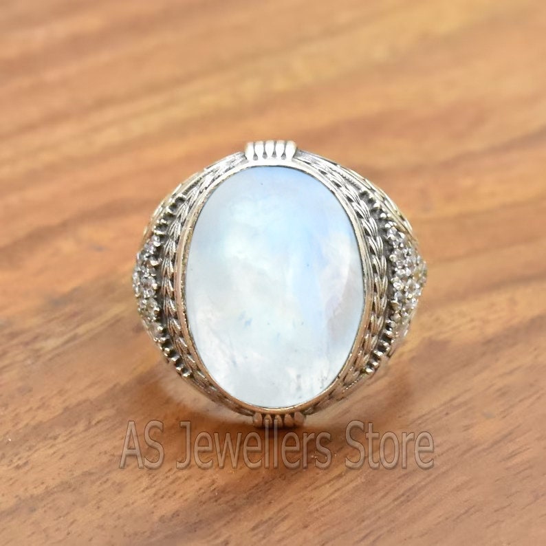 May include: A silver ring with a large oval moonstone set in a detailed silver band. The band is decorated with small silver beads and a rope-like design.