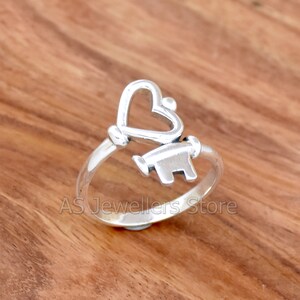 May include: A silver ring with a heart-shaped key design. The ring is on a wooden surface.