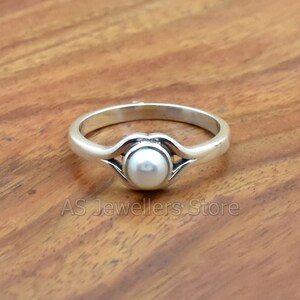 K&ouml;nnte beinhalten: Ein silberner Ring mit einem wei&szlig;en Perlmutt, das in einem zarten, geschwungenen Design gefasst ist.