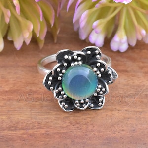May include: A silver ring with a large, flower-shaped setting. The petals are black with small silver dots, surrounding a large, round, iridescent blue-green stone. The ring is on a wooden surface, with blurred flowers in the background.