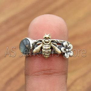 Pode incluir: Um anel de prata com um amuleto de abelha de ouro e uma pedra preciosa preta. O anel também tem um amuleto de flor de prata.
