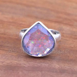 May include: A silver ring featuring a large, pear-shaped gemstone with a faceted surface. The gemstone displays a mix of purple, red, and green colors. The ring is set against a wooden background, with the text "AS Jeweller Store" visible.