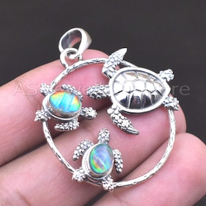 May include: A silver pendant featuring three sea turtles within a circular frame. Two turtles have iridescent opal-like shells, while the central turtle has a textured, silver shell. The pendant has a loop for a chain.