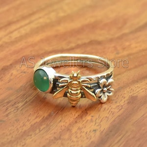 Pode incluir: Um anel de prata com uma gema verde, uma abelha dourada e duas flores prateadas. O anel está sobre uma superfície de madeira.