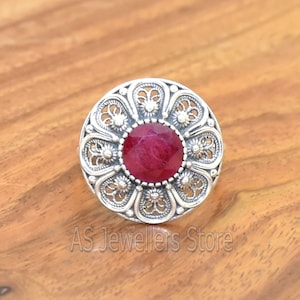 May include: A silver ring featuring a red gemstone set within a filigree design. The ring is round and has a floral pattern.
