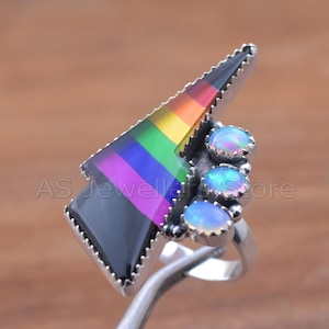 May include: A silver ring featuring a triangular design with a rainbow-colored inlay and a black base. The ring is accented with three oval, light blue opal stones. The ring has a unique, geometric design.