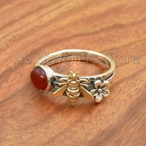 Pode incluir: Um anel de prata com uma pedra de cornalina vermelha, um amuleto de abelha de ouro e dois amuletos de flores de prata.