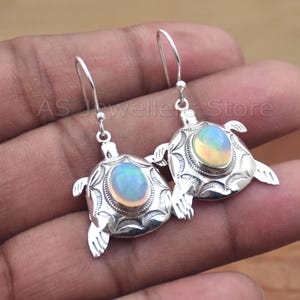 May include: Silver turtle earrings with iridescent oval-shaped stones. The earrings have a detailed design, with the text "AS Jewellers Store" visible in the background. The earrings are designed with a hook closure.
