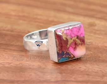 May include: A silver ring with a square pink, gold, and turquoise gemstone. The ring is set on a wooden surface.