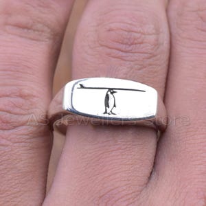 May include: A silver signet ring with a penguin and surfboard design. The penguin is black, under a black surfboard. The ring has a rectangular face and a polished finish. The ring is likely made of sterling silver.
