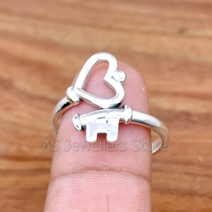 May include: A silver ring with a heart-shaped key design. The ring is on a finger.