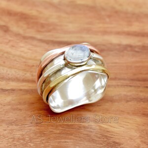May include: A silver spinner ring with a central, round, light blue gemstone. The ring has bands of silver, copper, and gold-coloured metal. The ring is displayed on a wooden surface.