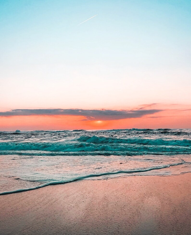 Peut inclure: Une vue panoramique d'une plage de sable avec des vagues turquoise de l'oc&eacute;an qui se brisent sur le rivage. Le ciel est un d&eacute;grad&eacute; vibrant de rose et de bleu avec une seule tra&icirc;n&eacute;e blanche d'un avion volant au-dessus.