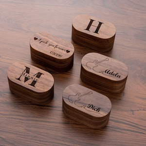 May include: Four wooden ring boxes with different engravings. One box has the letter "H" engraved on it. Another box has the letter "M" with the name "Minnie" engraved on it. The third box has a guitar and the name "Adela" engraved on it. The fourth box has a guitar and the name "Dick" engraved on it.