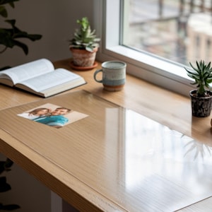 May include: A transparent desk protector rests on a light wood surface, with a photograph of a couple beneath it. A mug, open book, and potted plants are also on the desk, near a window. The scene suggests a workspace or home office setting.