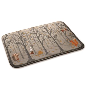 May include: Rectangular bath mat featuring a whimsical forest scene with bare trees, falling leaves, and woodland creatures. The design includes a hedgehog, rabbit, and fox. The mat has a neutral color palette with brown, gray, and orange accents.